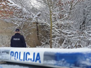 zimowa sceneria,policjant odwiedzający osoby narażone na wychłodzenie. na pierwszym planie radiowóz, w tle policjant idący w stronę zabudowań
